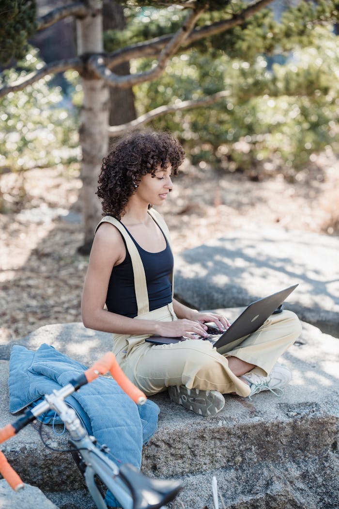Side view of young ethnic female remote worker with curly hair in casual sitting on stone with crossed legs and working on laptop in green park